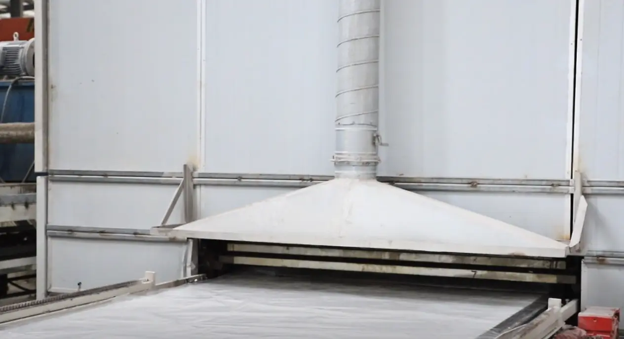 A conveyor belt carrying a quartz slab through a machine with an exhaust system in a manufacturing facility.